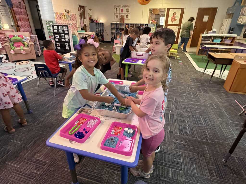 Kids in a classroom standing at a table with craft materials in front of them.