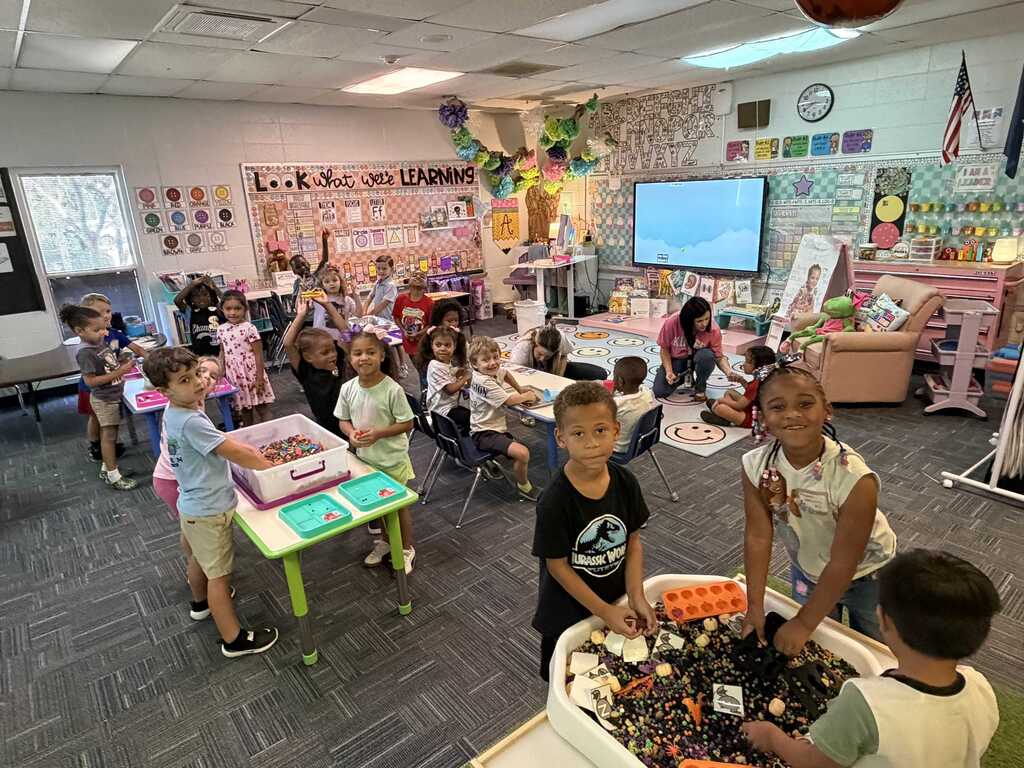 Children in a classroom stand around tables, one group with colorful beads, a monitor in the background.