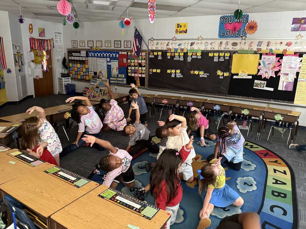 Group of children seated in a classroom with a teacher, engaging in exercise activities on a carpet.