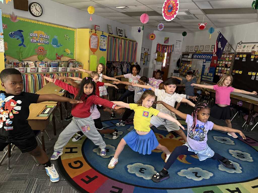 Group of young children in a classroom, dancing and stretching on a circle mat with alphabet letters.