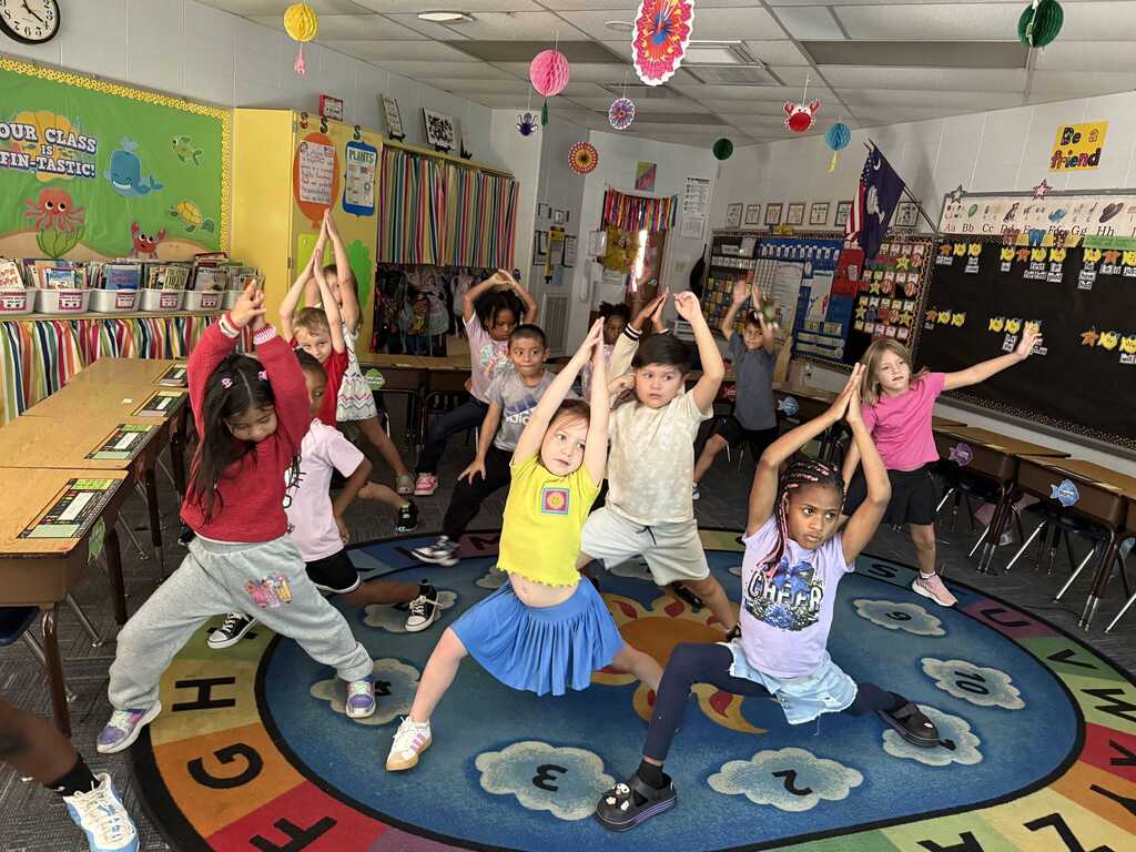 Group of children dancing in a colorful classroom with a round carpet, desks, and classroom materials.