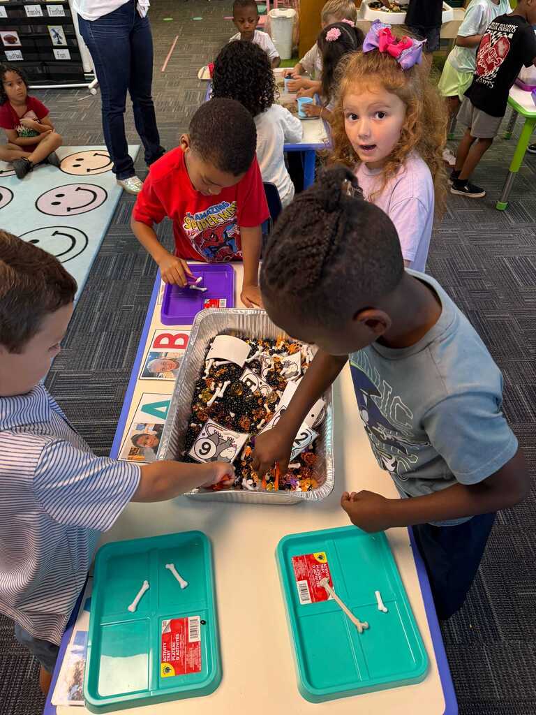 Four children are gathered around a table with food in a classroom setting. The children are interacting with each other.