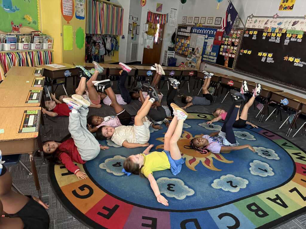 Children are performing stretches on a mat in a classroom. A colorful floor mat has letters and numbers.