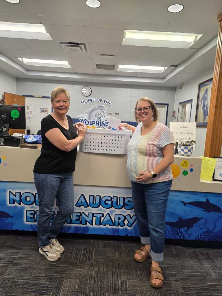 Two women stand behind a counter in a room. One holds a basket while the other smiles. Text "Nous Elementary" and an image of dolphins are on the counter.
