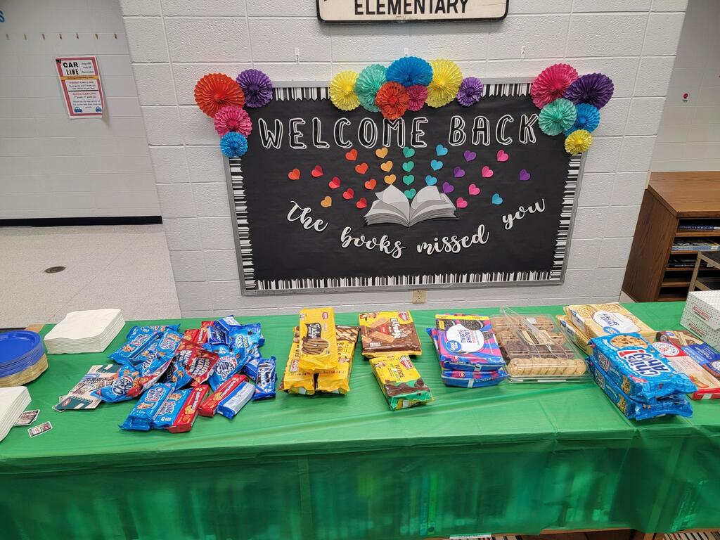 A table with a green tablecloth displays books, candies, and other items. Behind it, a blackboard reads "Welcome Back the books missed you".