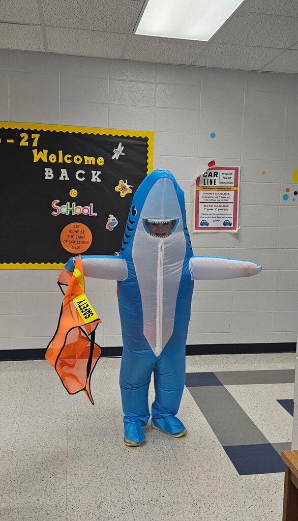 A person in a blue shark costume with an orange safety vest stands in a hallway. Behind is a bulletin board with "Welcome Back School" and other text.