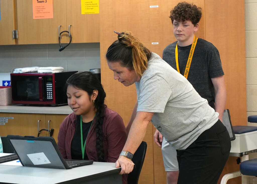 Two women in a room with cabinets, one looks at a laptop while the other looks over her shoulder.