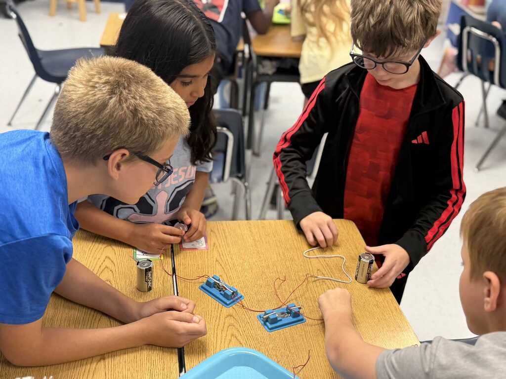 Four children are seated at a table, engaged in a science experiment. They are using batteries and wires to complete a circuit.