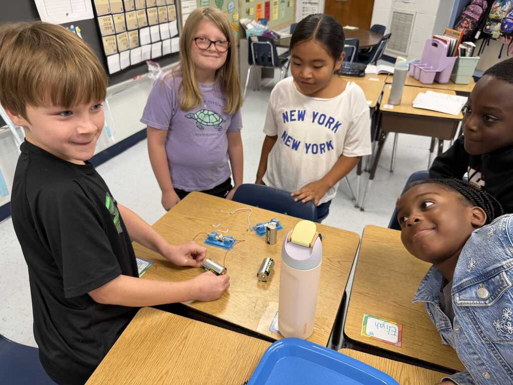 Five children in a classroom, one child sits, another leans over a desk with a science experiment.