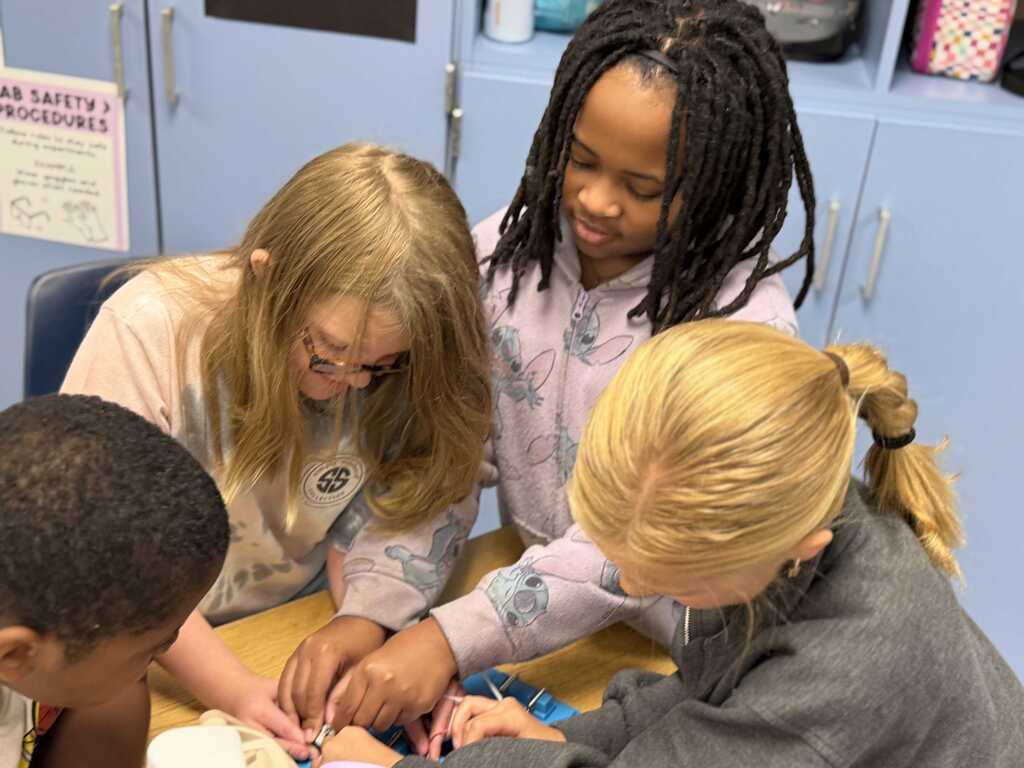 Three students with a teacher in a classroom, one student wearing glasses and two with ponytails.