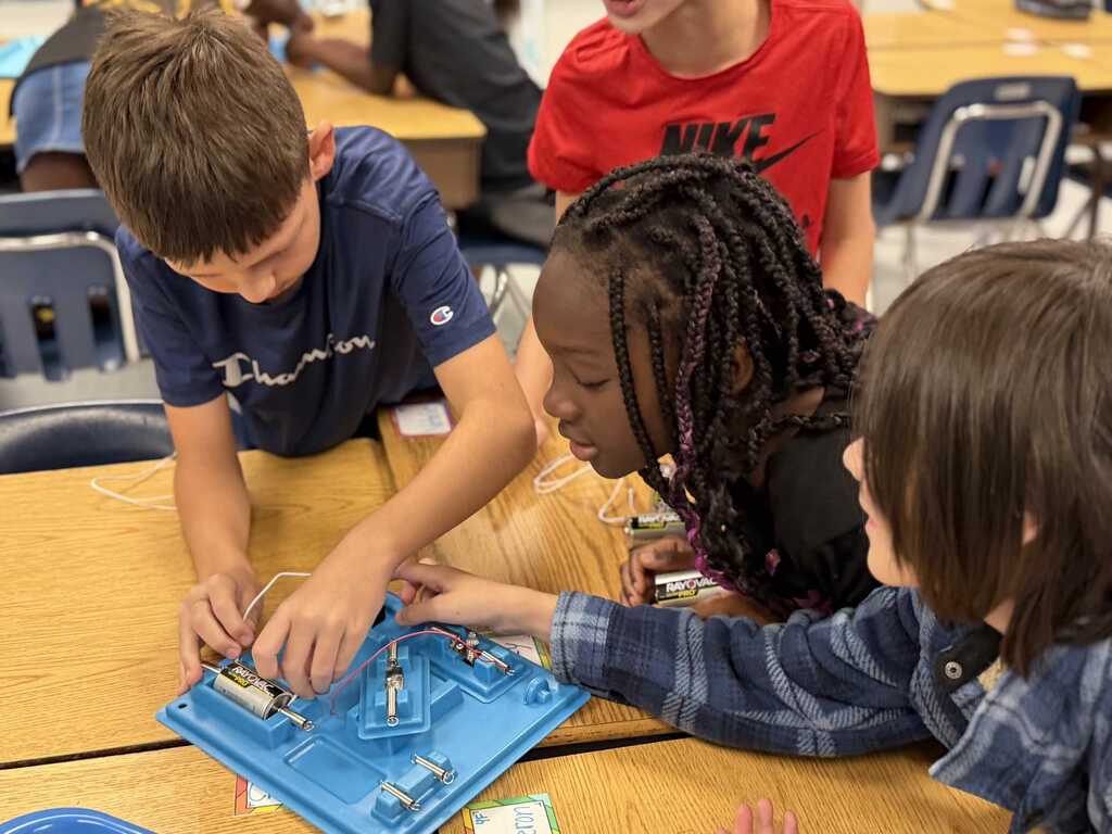 Three children at a table, one boy and two girls, working together on a blue electronic circuit board.