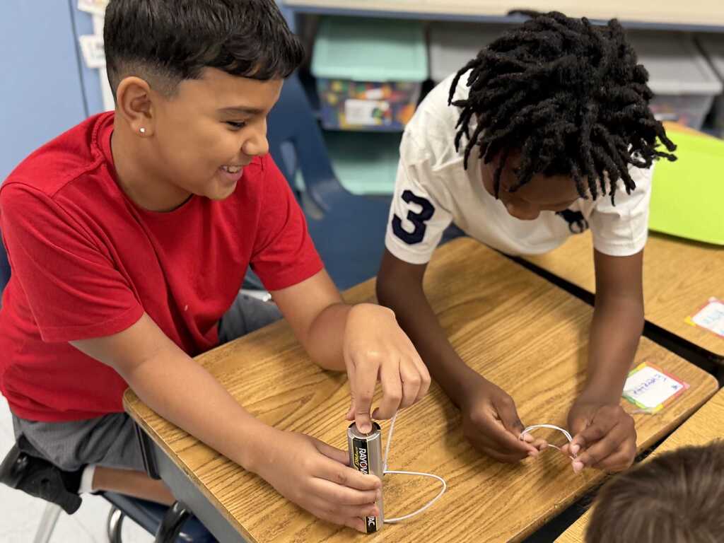 Two young people seated at a desk are working together on a science project involving wires and a battery.