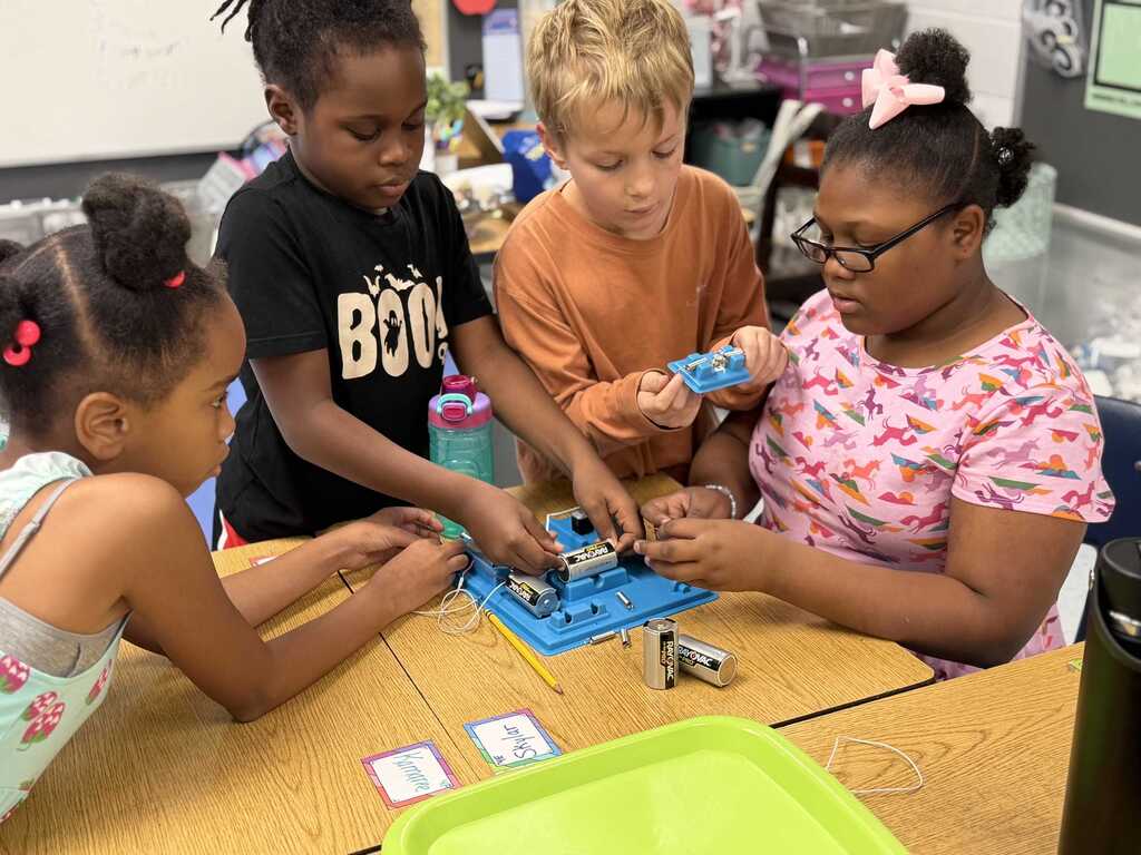 Four children work on a small electronic device on a desk. One child wears glasses.