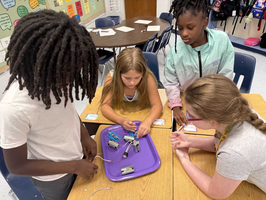 Four students are seated around a table, working on a project. One student is holding a purple tray with tools.