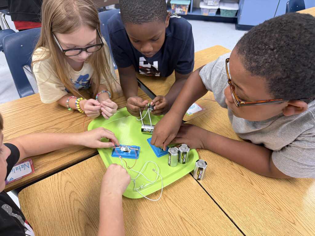 Four children wearing glasses are seated at a table, focusing on a green board with electronic components.