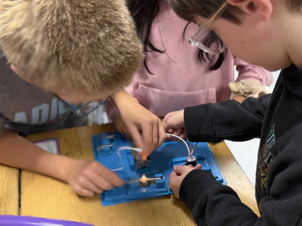 Two children work on an electronic project, with one holding a blue board and the other adjusting a wire.