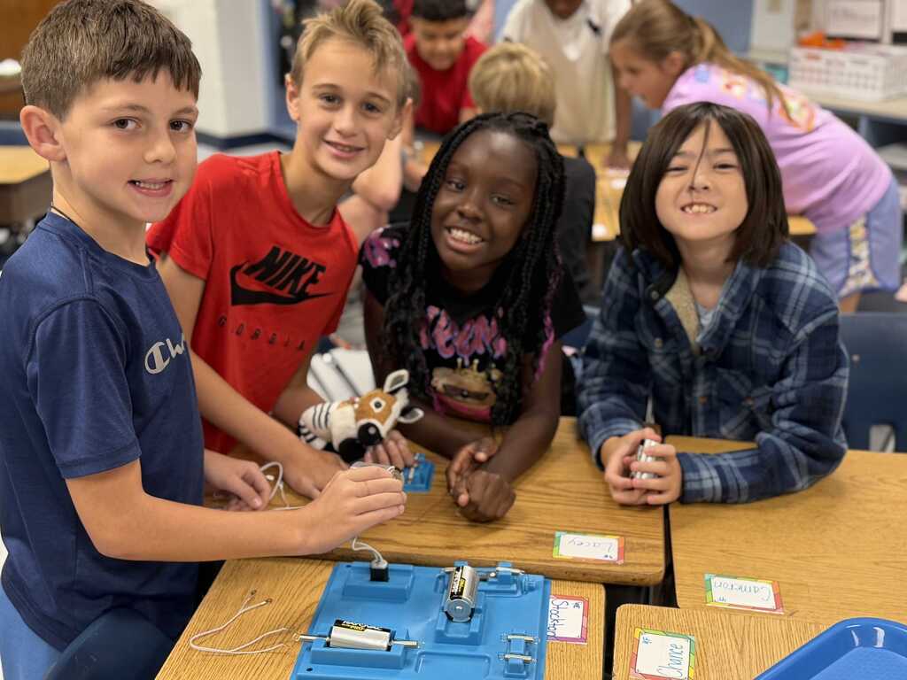 Five children, two girls and three boys, seated around a table with electronic parts. Background includes a classroom setting.