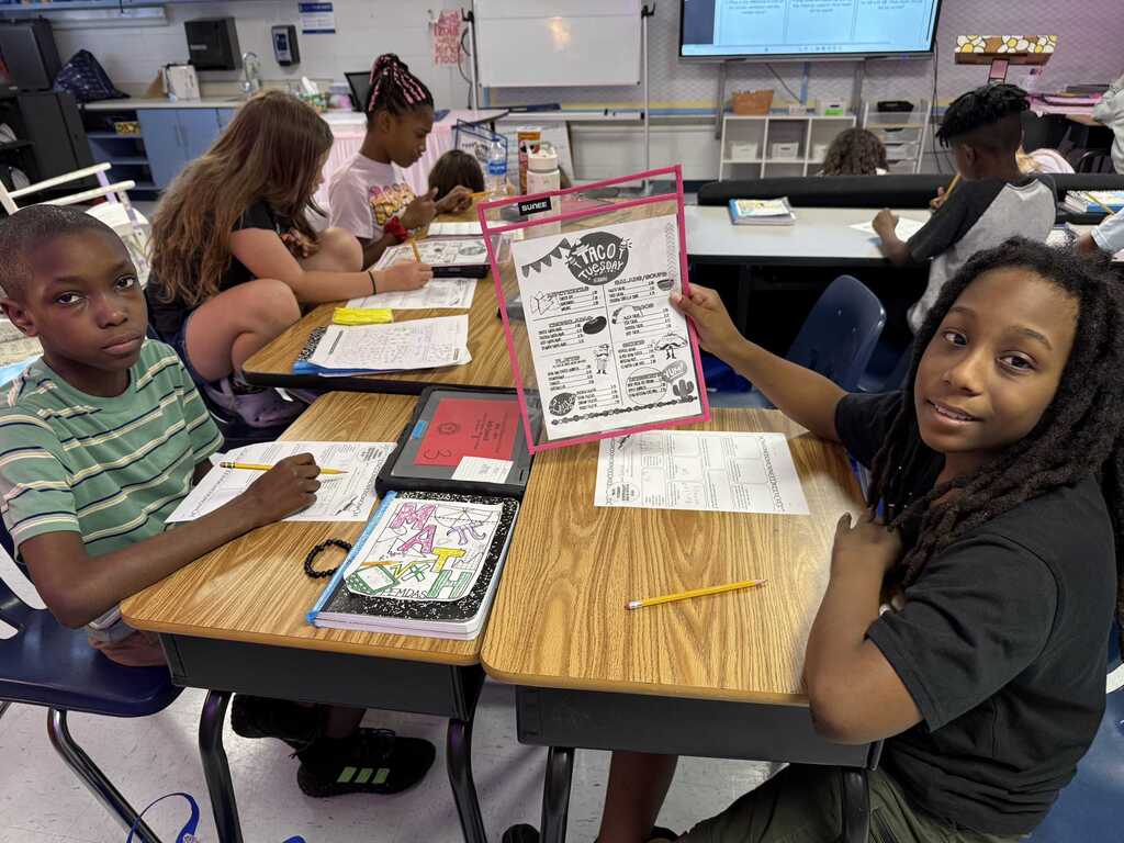 A classroom with students sitting at desks. One student holds a colorful sheet of paper while another looks on.