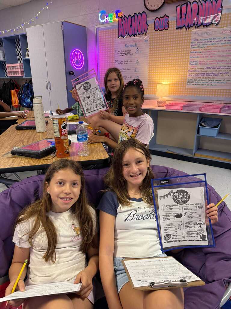 Three girls sit on a purple bean bag in a classroom. Two hold papers, one holds a folder. Behind them, a table has books and a clock.