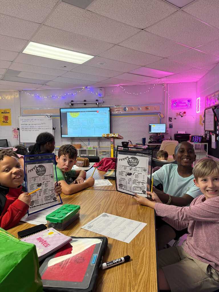 Children seated around a desk with papers, holding up drawings. Bright lights on the ceiling. Behind them is a screen.