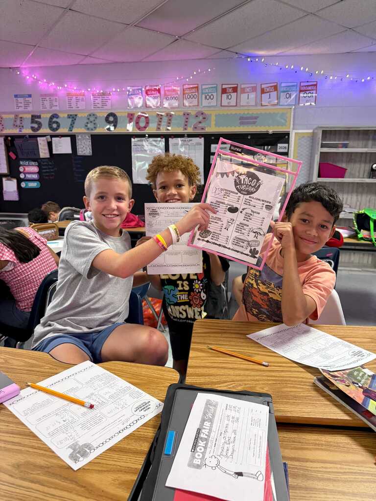 Three kids seated at a table, holding up paper, with school materials in a classroom with a white ceiling.
