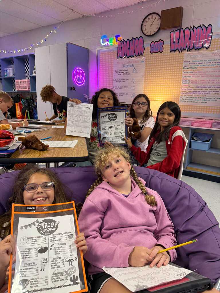 Students sitting in a classroom holding papers. One holds a paper with the word "Taco".