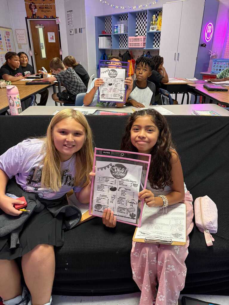 Two girls sit on a couch holding papers with printed text and images, smiling. Behind them, a classroom with desks and students is visible.
