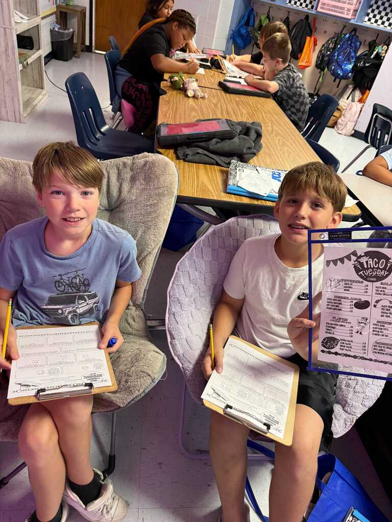 Two boys seated in chairs, holding papers and clipboards. One holds a clipboard with text. Behind them, a table with papers, and other students seated at tables.