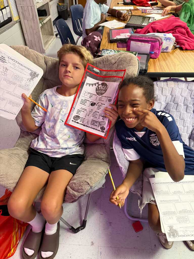 Two children sitting on chairs in a classroom. One holds a paper, and the other holds a pencil.