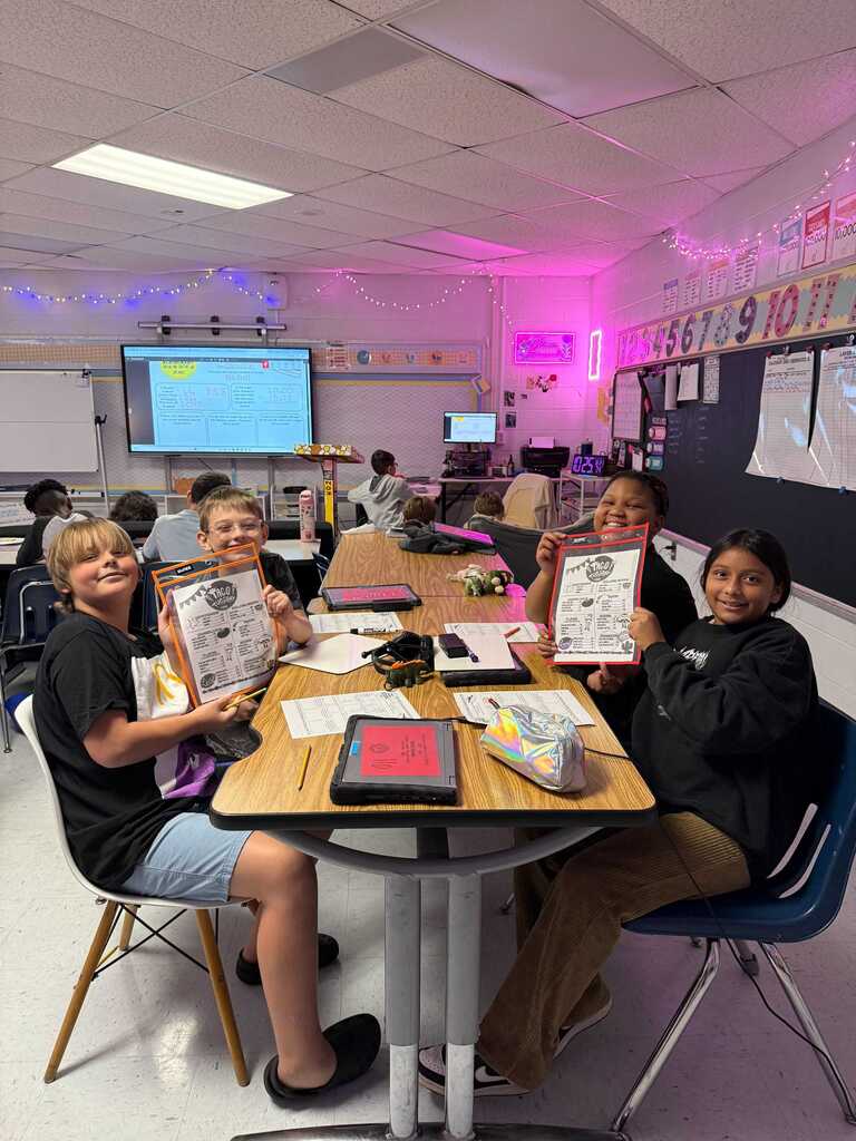 Four children in a classroom with a table, holding up papers, smiling. A monitor is visible in the background.