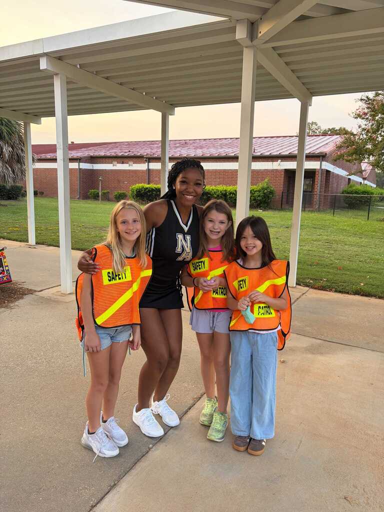 Four girls in matching orange vests and sneakers, one holding a water bottle, standing under a shelter. Behind them, a grassy area with a building.