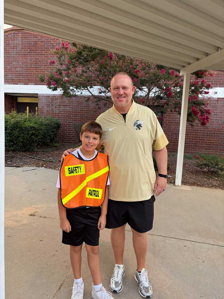 Man and a child stand outside a building. Man wears a yellow shirt, black shorts, and white sneakers. Child wears an orange vest, black shorts, and white sneakers.