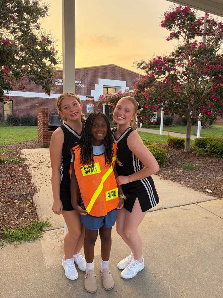 Three people in matching uniforms pose outside. One wears an orange vest. Behind them is a building with a lawn and trees.