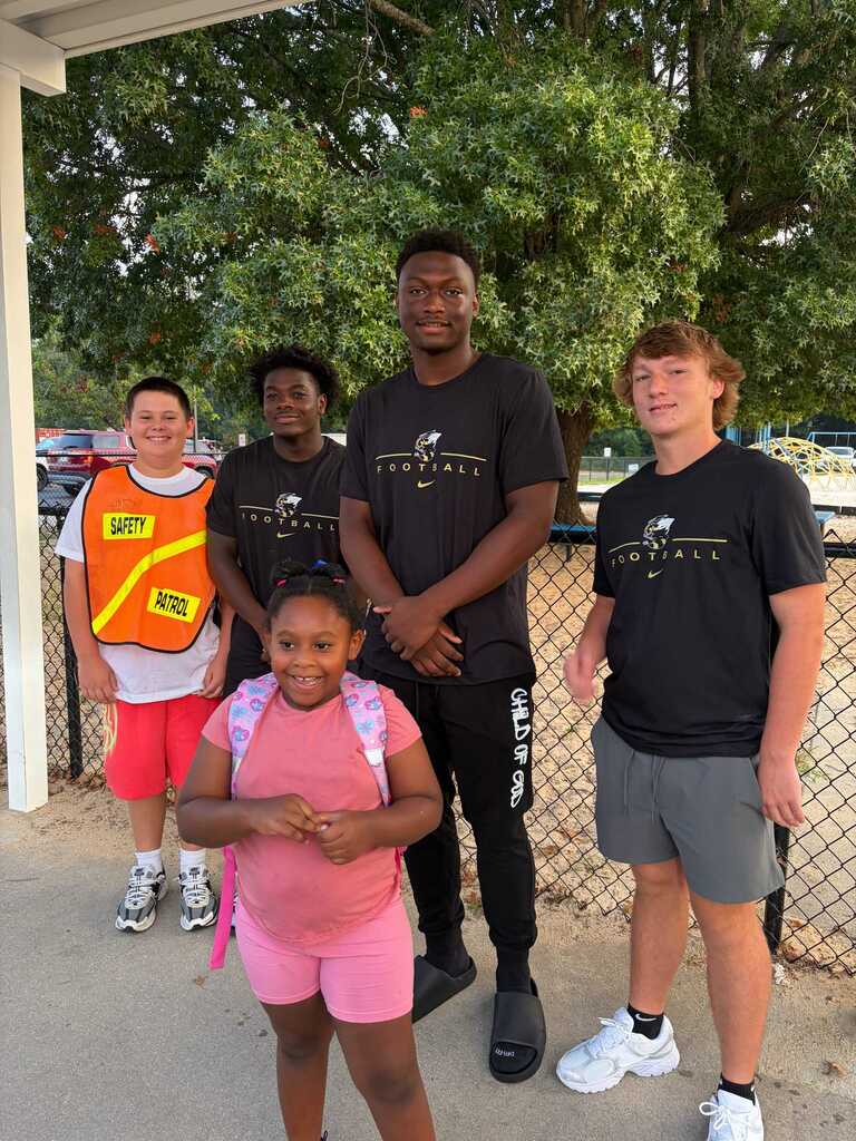 Five individuals stand near a fence in a park. A child with a backpack is in the center. Tall men in black shirts are on either side.