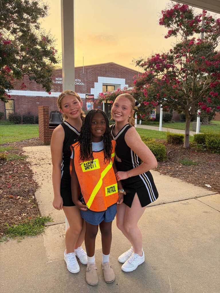 Three individuals pose outside on a pathway, two with uniforms and one in a safety vest. Behind them, a building is visible.