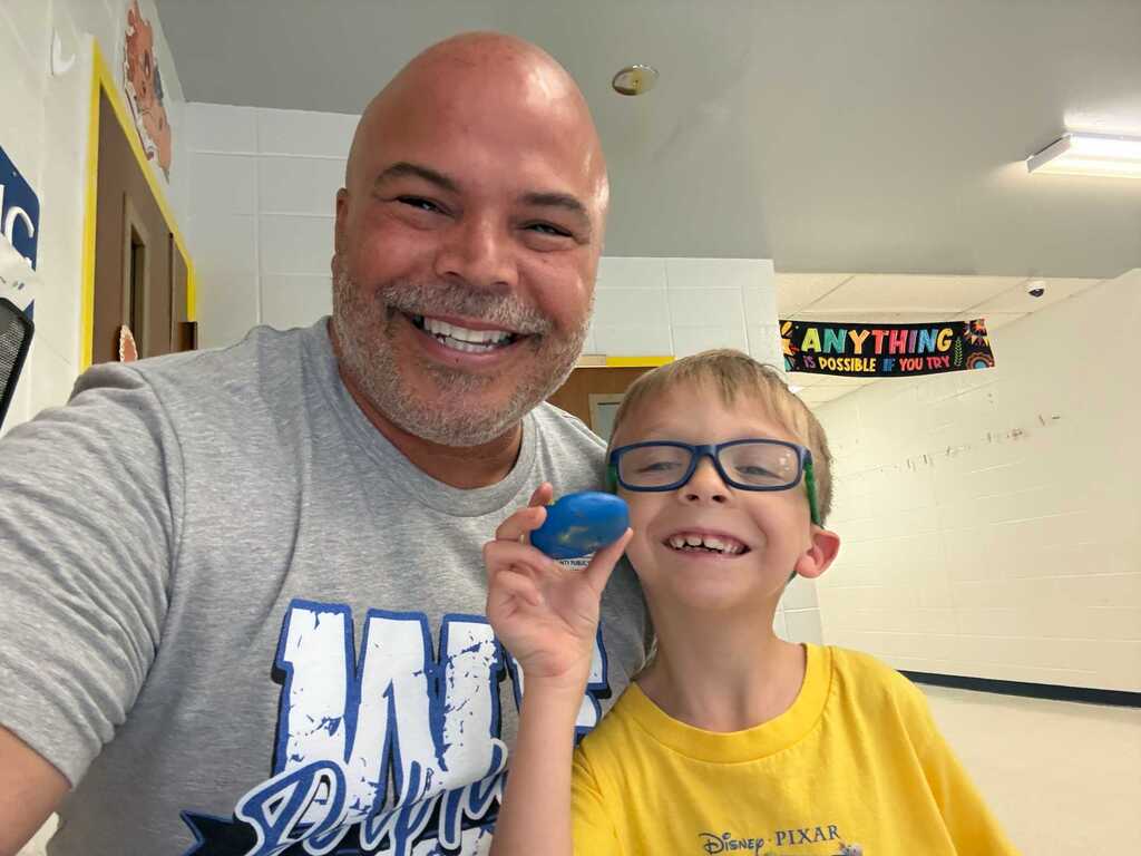 A smiling man in a gray t-shirt and a boy in a yellow shirt stand together. The boy holds a blue object and wears glasses. They are inside a room with white walls.