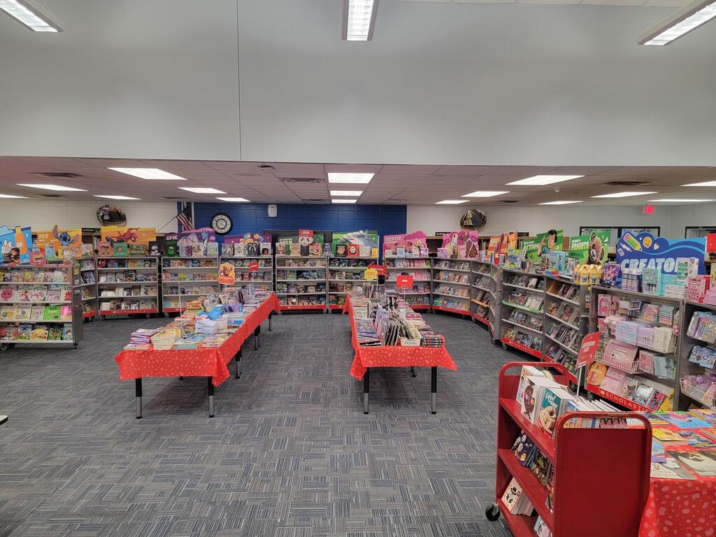 A bookstore with tables, bookshelves, and books. Ceiling lights and a clock are visible. The floor is carpeted.