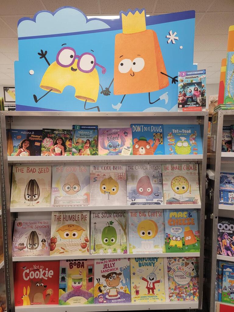 Bookshelf with books and a colorful character sign above. Titles include "The Cookie," "The Egg," "The Fish," and more.