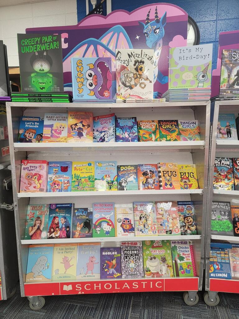 Display of colorful children's books on a white shelving unit with a red wheel cart labeled "Scholastic."