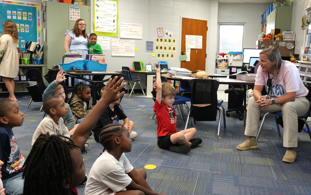 Kids sit on the floor in a classroom with raised hands; a woman sits nearby on a chair.