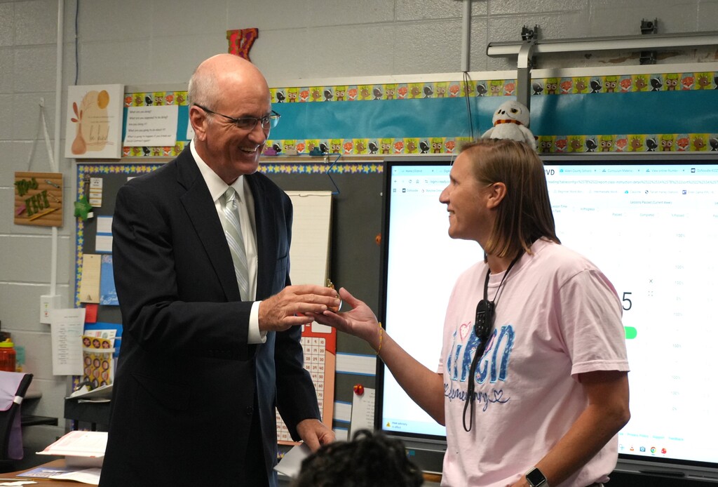 A man in a suit and a woman in a pink shirt interact in a classroom. Behind them, two monitors display information.