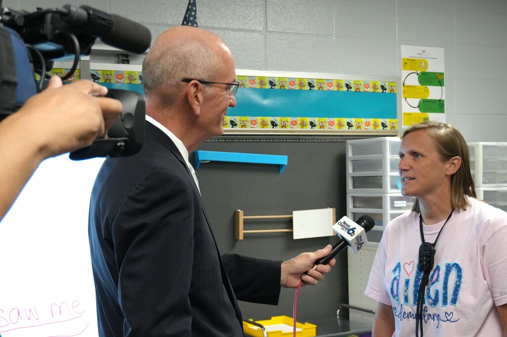 A man in a suit interviews a woman in a white shirt. They are in a room with a whiteboard, shelves, and a camera.