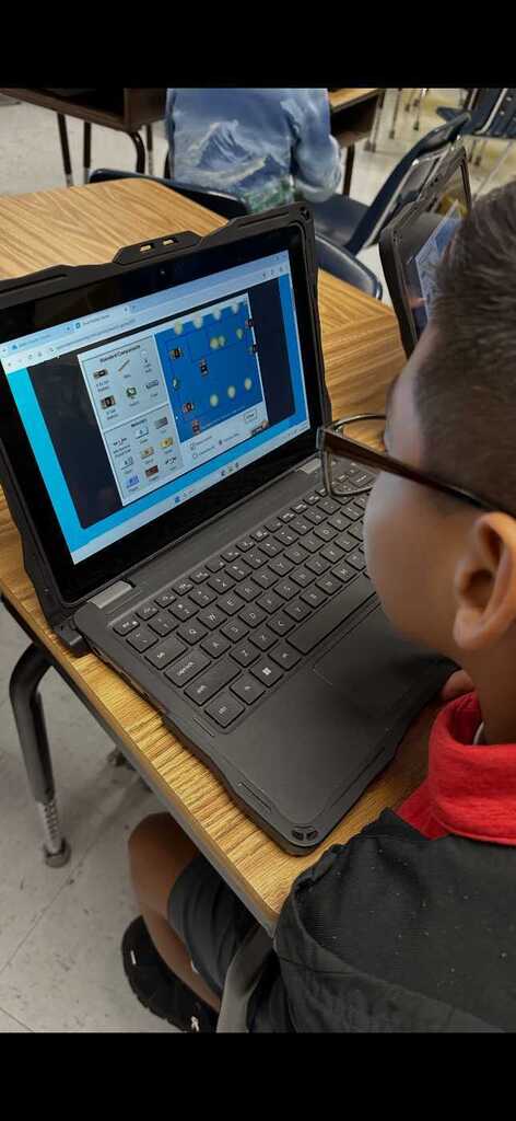 A boy wearing glasses sits at a desk using a laptop, screen showing a blue grid.