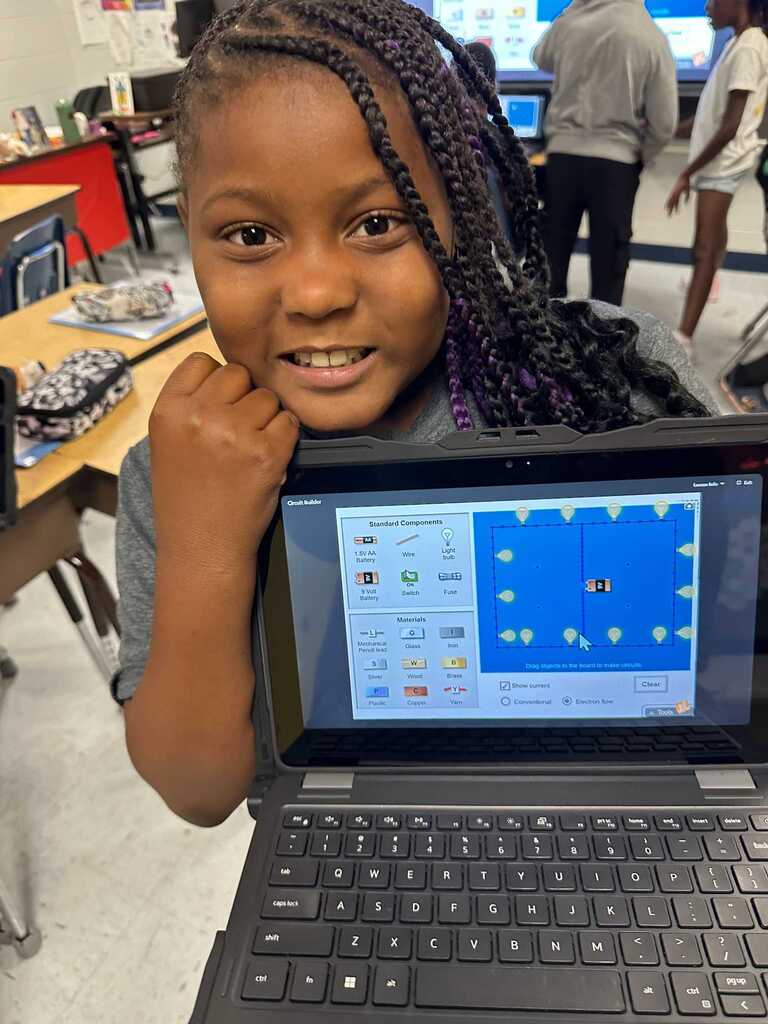 A young girl, smiling, holds a laptop showing a game on the screen. Behind her, a classroom with desks, backpacks, and other students is visible.