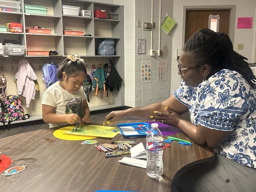 Woman and child at a table in a classroom, with crafts, papers, and a water bottle.