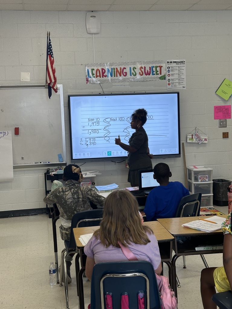 A classroom with a teacher in front of a projector screen. Students sit at desks with laptops.