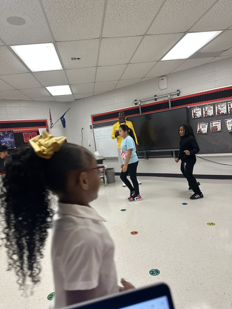 Children in a gym play a game on the floor, colored circles scattered. Two adults in yellow and blue shirts oversee.