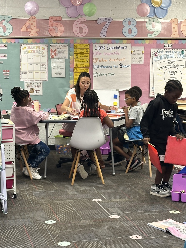 Classroom with students and teacher. Teacher helps young children at a table. Bright colors and balloons.