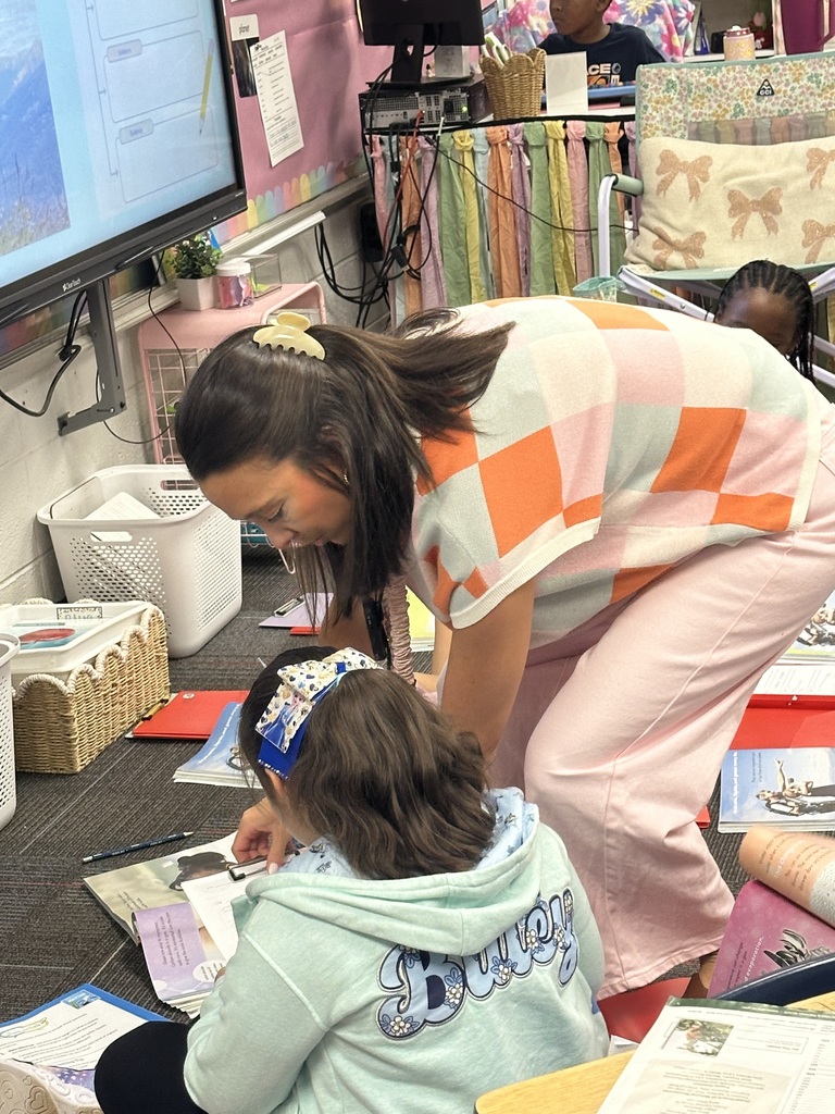 Woman in pink and white patterned top and pants assisting a girl with a green hoodie, both on floor.