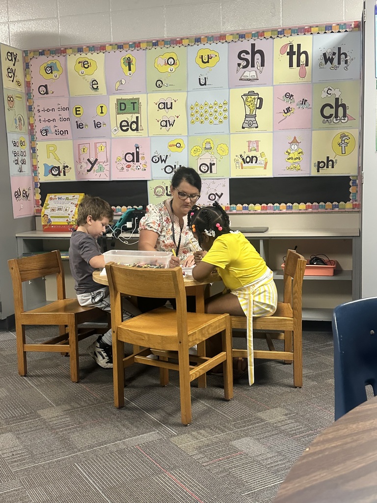 A classroom with a colorful wall. A woman and two children are seated at a table, working on a project.